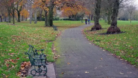 StephenMcKay/Geograph Moor Park, a flat grassy park with a wide asphalt path through autumnal trees. There is a green metal bench to the left foreground.