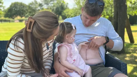 Rory lifts his top to show Harriet the scar on his tummy. She is looking at the scar. Her mother Emily sits next to them.