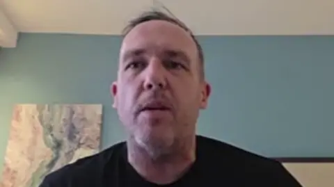 The image shows a pale faced man with facial stubble wearing a dark shirt, seated indoors against a background with light‑coloured walls. A piece of framed artwork with abstract patterns hangs on the wall to the left.