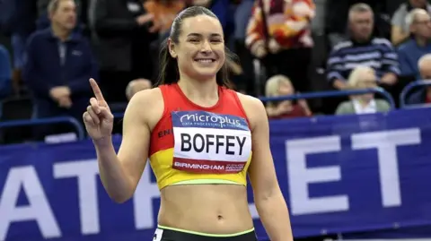A female athlete smiling with a finger in the air. She has dark brown hair that is tied back. She is wearing a red and yellow vest with her name on the front. The background behind her is blurred but we can see a crowd of spectators in the stand. 