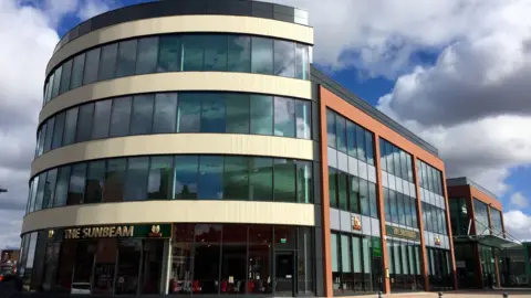 A large office building with a curved edge with a wall of windows, and a frontage with orange accents and windows. A gold sign reads "the sunbeam"