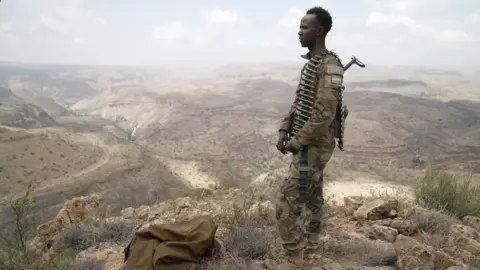 BBC/Amensisa Ifa A young man in camouflage clothing stands on the right with a belt of bullets draped over his shoulder. He is looking out over a valley in a remote mountainous area.