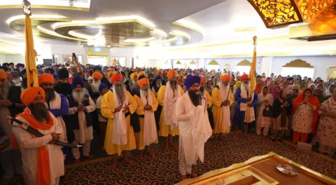 Dozens of male Sikhs in traditional dress gather inside the temple with a large crowd of men and woman behind them. The men have yellow, red, blue and orange turbans on, some holding flags. There is a gold design on the ceiling and walls.