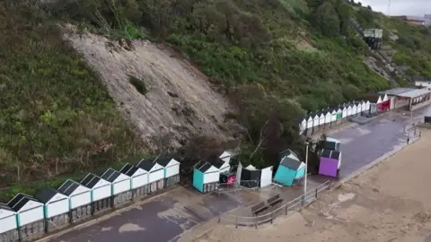 Cluster of beach huts several with turquoise sides and one with purple with tress and debris collapsed from cliffs behind onto them.