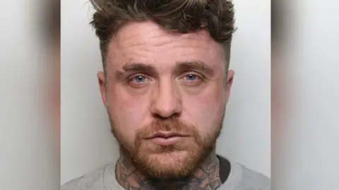 A police mugshot of Shane Davis. He is standing against a light-coloured background. He has brown hair and a beard, with tattoos around his neck.