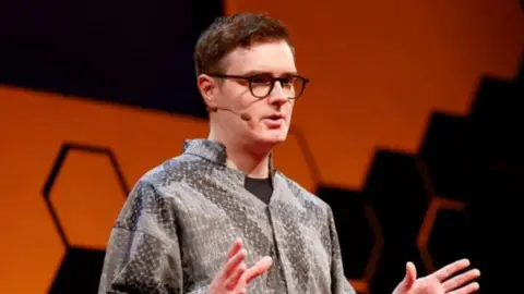 Welsh computer scientist Llion jones stands on stage at a TED-AI conference in San Francisco, wearing a grey patterned, loose-fitting shirt. The background features a bold orange shape with a honeycomb-style black geometric design. Llion wears glasses and a microphone and is mid-presentation, with both hands raised in a gesturing position.