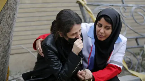 Anadolu via Getty Images A woman consoles another woman who cries while sitting on the side of the street in Tehran on Monday.