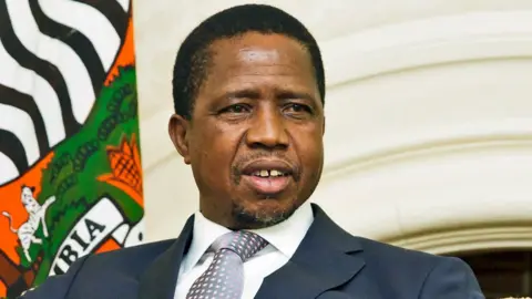 Edgar Lungu, in a suit and tie, sits next to a Zambian flag