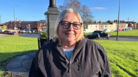 blonde woman with mid-length hair and black framed glasses wears a black rain jacket and smiles. In the background is a village green with the black lampost. Behind are residential houses.