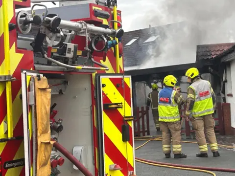 Fire crews outside a house with smoke billowing from its roof