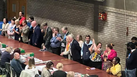 Finn Cullum/BBC A row of standing men and women, some with blue rosettes and some with yellow rosettes on one side of a series of tables put together to make one long table. On the other side are people sitting down with ballots in front of them. They are in a hall in Huntingdonshire. 