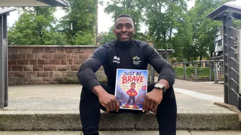Joel Fearon, who is dressed in sports gear is sat on a stone step, with a wall and trees behind him. He is smiling at the camera and holding up his book, which is called Just Be Brave.