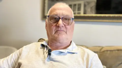 Alex Dunlop/BBC A bald man wearing glasses and a grey top with a small black microphone attached to the collar sits on a cream sofa. He is looking at the camera and is not smiling. 