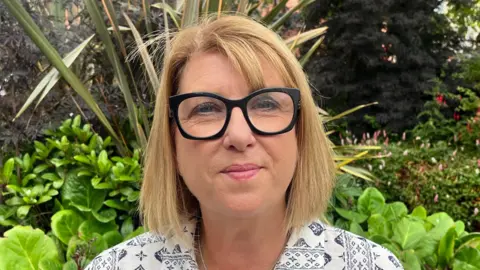 A woman with chin-length blonde hair stares at the camera as she wears black glasses and pink lipstick. She is wearing a white shirt with a black pattern on it. Behind her is flowerbed with green plants in it.