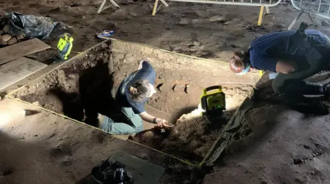 Archaeologists at a site are digging in a rectangular hole in the ground surrounded by metal fences. They are wearing head torches