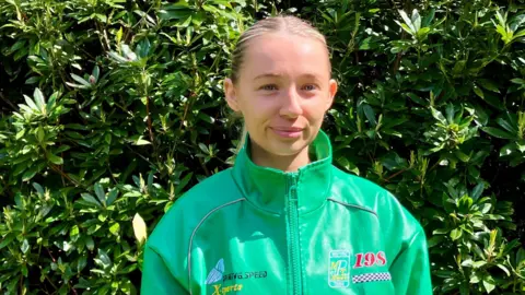 Amie Donaghy has blonde hair tied back in a ponytail and is wearing a light green driving-style jacket with a high collar. She is standing in front of a hedge.