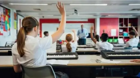 a small class of children raise their hands as a teacher stands facing them in front of a whiteboard, holding what looks to be a piece of paper