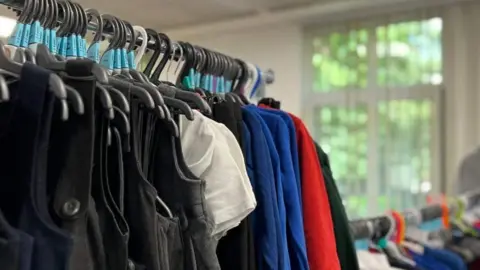 A rail of uniforms with white shirts, blue jumpers, grey pinafores and red sweatshirts on hangers.