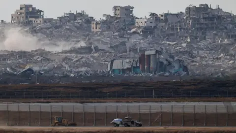 An Israeli military vehicle moves near the Israel-Gaza border. In the background, there is a mass of debris and smoke from destroyed buildings in Gaza.