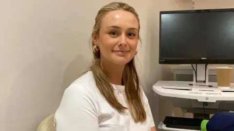 Jade Linden has long blonde hair and is wearing a white, short-sleeved t-shirt and gold hoops. She is is smiling at the camera and sitting by a wall, with a computer screen to the left of her.