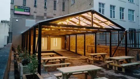 Tom Arber A number of picnic benches sit beneath a metal canopy at the front of a brick building. On the wall is a sign saying Loading Bay.