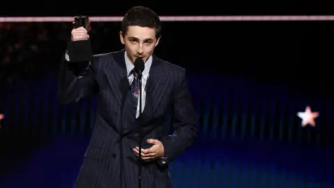 Timothée Chalamet, who is wearing a pin-striped suit, white shirt and patterned tie, accepts the Best Actor Award for Marty Supreme at the 31st Annual Critics Choice Awards in Santa Monica, California. He is holding his award in the air as he talks into a microphone. 