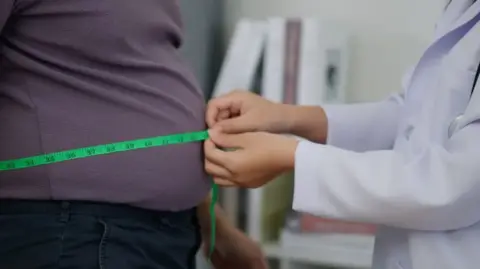 A tape measure is held around a man's waist, over his grey top, by a woman in a medic's white coat