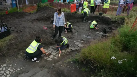 BBC Image shows people in high-visibility tabards digging into the earth, uncovering what look like sections of cobbled streets below the soil