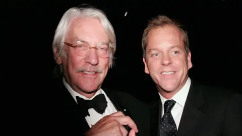 Actors Donald Sutherland (l) and Kiefer Sutherland (r) wearing black suits with white shirts. Donald has white hair and glasses and a moustache; Kiefer is clean-shaven with cropped hair and is looking down, both are smiling 