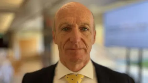 Alex Dunlop/BBC Kevin Keable was in the middle of the image looking towards the camera. He had short grey hair and was wearing a white shirt, with a black suit jacket and yellow tie.