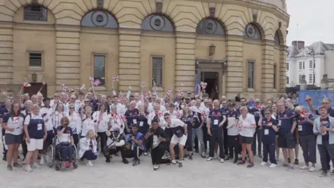 A large group of athletes waving British flags and stood outside the Sheldonian theatre in Oxford.