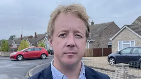 A head and shoulders image of Paul Bristow standing on a residential road, with bungalows in the background, a black car parked in a driveway and red car parked by a kerb.