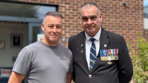 Two men are standing next to each other with their hands behind their backs. The man on the left is wearing a grey t-shirt. The man on the right is wearing a black blazer, tie and badges.