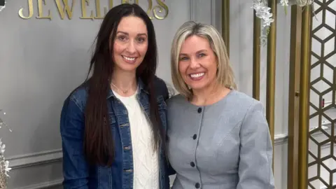 Two women pose for a photograph, smiling warmly, in front of a sign that says "Jewellers". The woman on the left has long dark hair and wears a dark blue denim jacket. The woman on the right has blonde hair and is wearing a grey top. 
