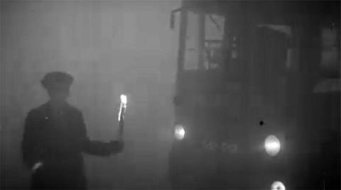 Black and white image of a uniformed official (perhaps a Bus conductor or policeman) guides a bus through the smog holding a flaming torch on the left hand side.  A bus on the right is following him.