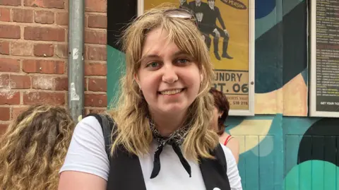 Chloe Aslett/BBC A woman with blond shoulder length hair. She wears a white t-shirt, black cotton tux, and black bandana around her neck.