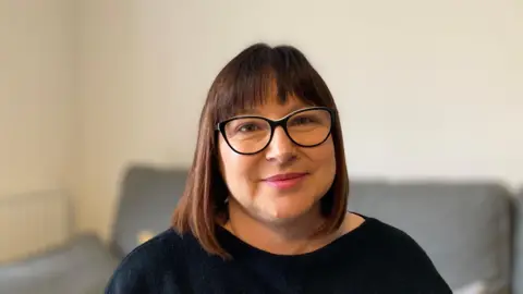 A woman with dark brown hair, black glasses and black top smiles at the camera. A grey sofa is slightly blurred in the background behind her.