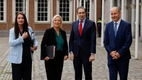 Reuters Little-Pengelly, left, has long brown hair and is wearing a light blue blazer. She is standing next to O'Neill, who has blonde hair tied back and is wearing a green blouse and black suit. Harris is next wearing a navy suit, blue shirt and red tie. He has grey hair. Martin is on the right, wearing a navy suit, blue tie and white shirt.