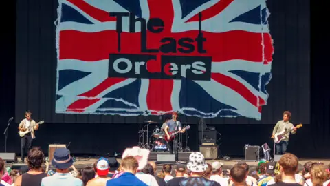 A band on stage in front of a Union Jack which says THE LAST ORDERS