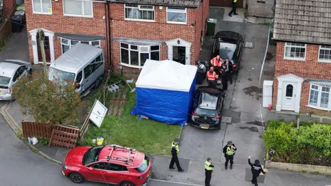 Police gather in front of a row of semi-detached properties. One house has a blue police tent in the front garden and has the door missing. 