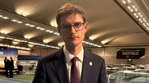 BBC/Simon Thake A man with short brown hair and glasses is wearing a dark suit, white shirt and burgundy tie. He looks incredibly sad.