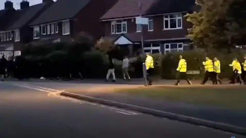 Other People dressed in black and police in yellow vests walking down a residential street. There are houses visible in the background