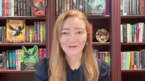 A woman with long blonde hair sits in front of a book case full of the Skandar book series that she has written. The books spines are all different colours with the pictures of the creatures that feature in them on them.