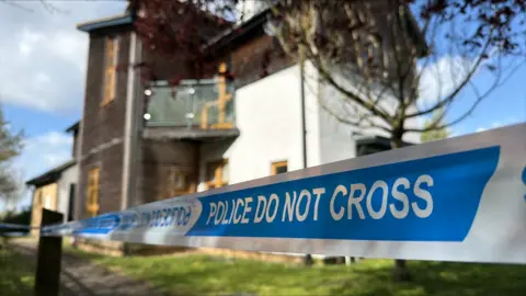 Blue and white tape stating POLICE DO NOT CROSS stretches across the front of the image. Behind is a blurred background of a white house with dark wooden cladding.