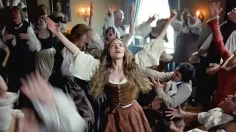 Searchlight Pictures Amanda Seyfried playing Ann Lee in a scene from the film The Testament of Ann Lee. She has long blond crimped hair and is wearing a brown suede corset and long brown skirt as she dances wildly along with men and women around her during Shaker worship.