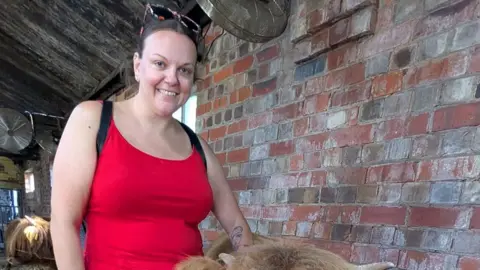 A photograph of Stacey Waring wearing a red top standing in a barn. She is by herself wearing dark sunglasses on her head and smiling at the camera.