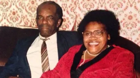 Ms Baton's parents sitting on a brown chair, and Mr Banton has his arm around Ms Burrell. He is wearing a blue suit jacket, white shirt and striped tie. He has very short black hair. Ms Burrell is wearing a red jacket and white pearl necklace, and has short black hair.