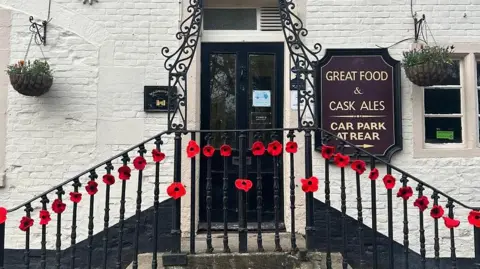 Holt Residents Front of a pub - black door in the middle with steps up to either side, poppies going up and down the railings.