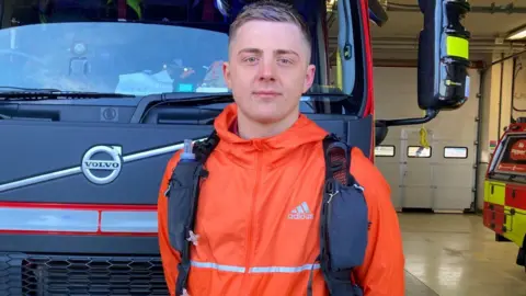 A man looks at the camera. He has an orange jacket on and has a bag over his shoulders. He is standing in front of a fire engine which is inside a station. 