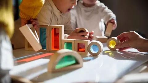 Three toddlers play with coloured shapes around a table at a nursery.